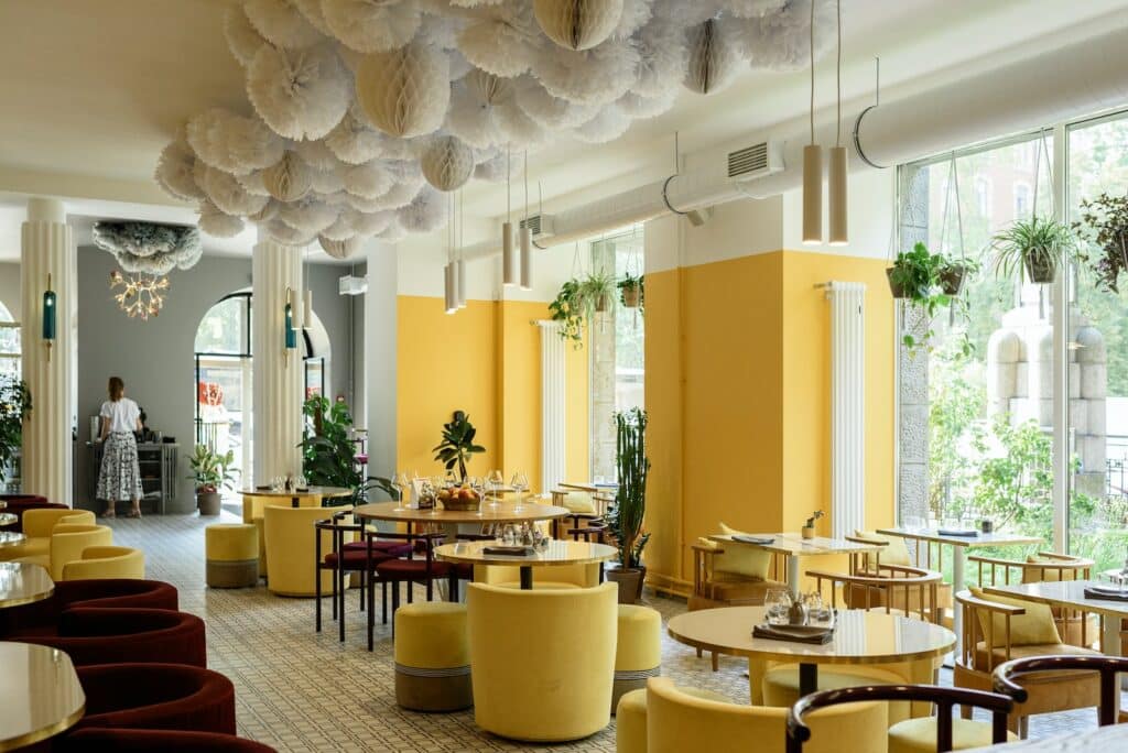 Creative interior design of modern spacious cafe with bright yellow walls and armchairs decorated with green plants and cozy lamps with big windows against lush greenery